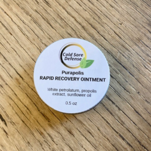 Purapolis Cold Sore Defense Rapid Recovery Ointment in an aluminum tin on a wooden table, made with white petrolatum, propolis extract, and sunflower oil for soothing cold sore relief.